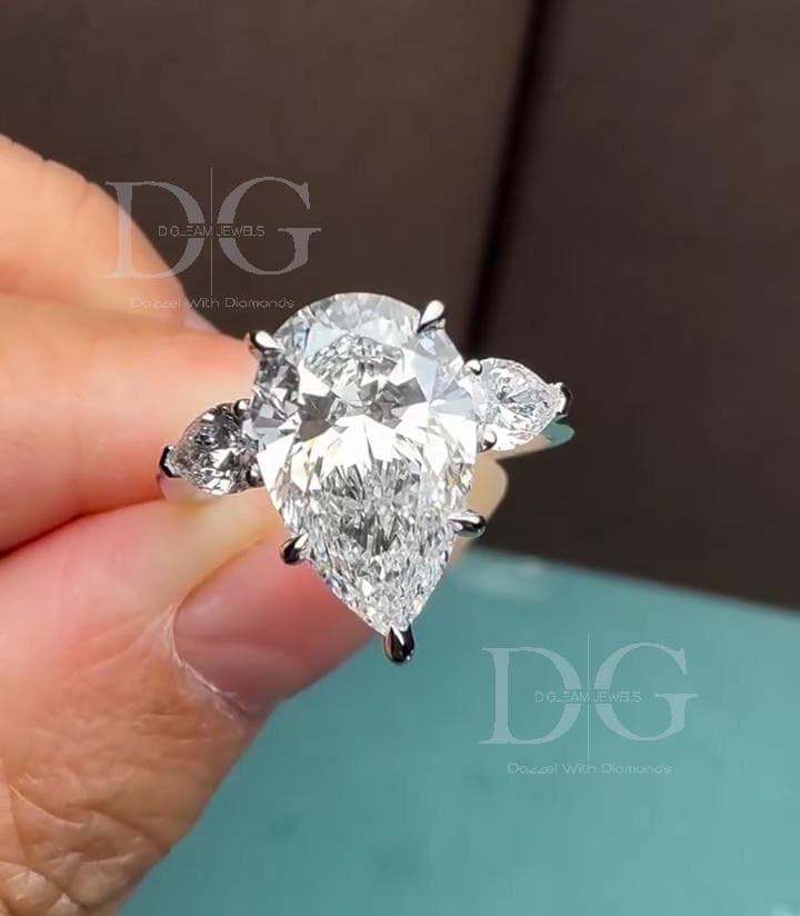 May include: A pear-shaped diamond engagement ring with three stones. The large, central diamond is pear-cut and flanked by two smaller, round diamonds. The ring has a silver band and is held against a blurred background.
