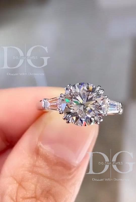 May include: A diamond engagement ring with a large, round-cut center stone and two smaller baguette-cut side stones. The ring has a platinum or white gold band. The center stone is surrounded by a halo of smaller diamonds. The ring is held up against a blurred background.