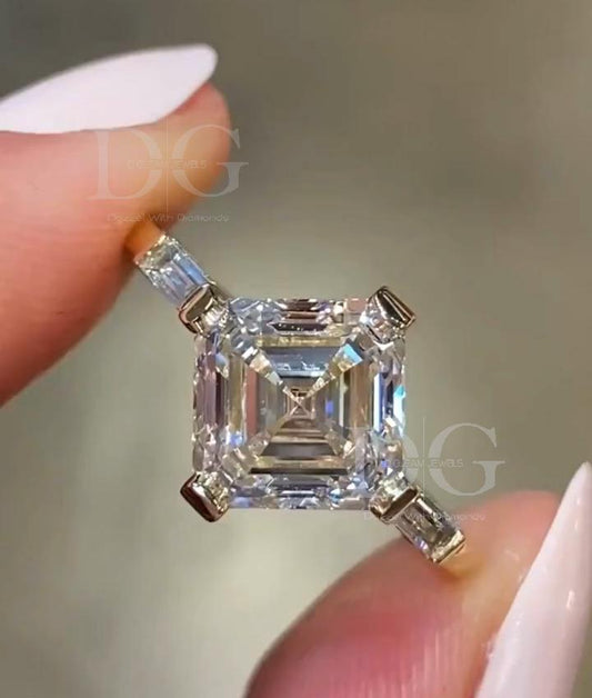 May include: A square-cut diamond engagement ring held between two fingers. The ring features a large, clear, square-shaped center stone with a multi-faceted design. Four smaller, rectangular diamonds are set on the corners of the square center stone. The ring's setting appears to be gold-colored.