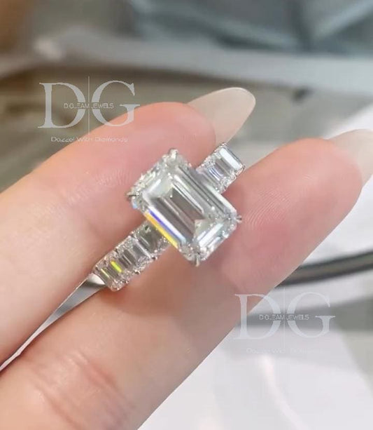May include: A diamond ring with an emerald-cut center stone and a band of smaller diamonds. The ring is set in a white metal, and the diamonds sparkle. The ring is held up against a light background. The ring is a piece of jewelry.