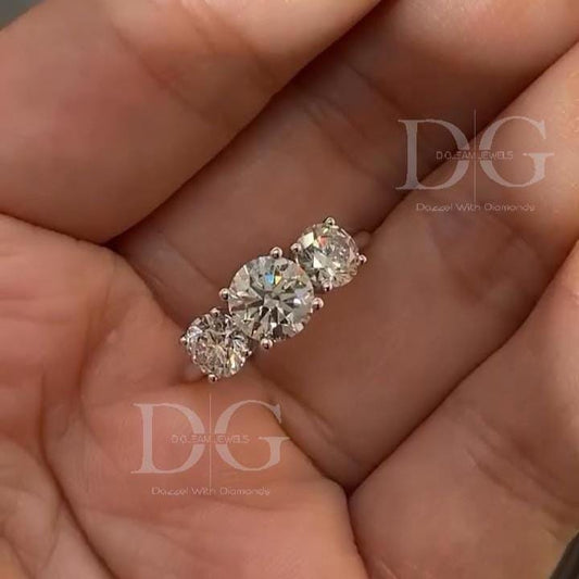 May include: A three-stone ring with a platinum band. The ring features three round-cut diamonds of varying sizes, with the largest diamond in the center. The diamonds are set in a classic prong setting, and the ring is displayed against a hand.