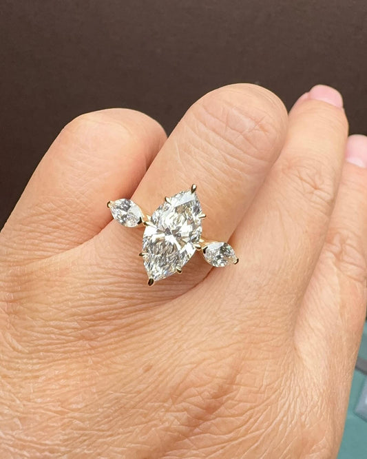 May include: A marquise-cut diamond ring with a gold band. The ring features a large, central marquise-cut diamond and two smaller pear-shaped diamonds on either side. The diamonds are clear and sparkling, set in a gold band. The ring is displayed on a hand.