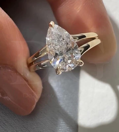 May include: A pear-shaped diamond engagement ring with a split shank band. The diamond is clear and faceted, set with prongs. The band is a polished gold color, and the ring is held up against a blurred background.