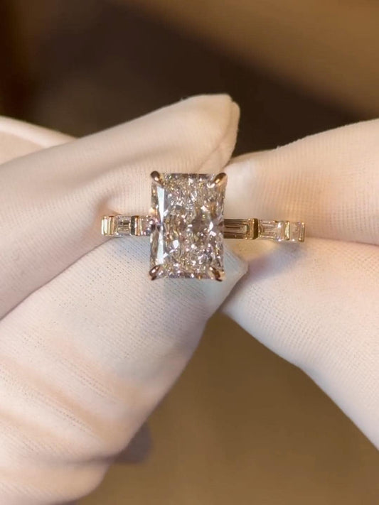 May include: A radiant-cut diamond engagement ring. The ring features a large, rectangular-shaped diamond as the center stone, with smaller baguette diamonds on either side of the main stone and on the band. The setting appears to be rose gold, with a delicate and elegant design, showcasing the brilliance of the diamonds.