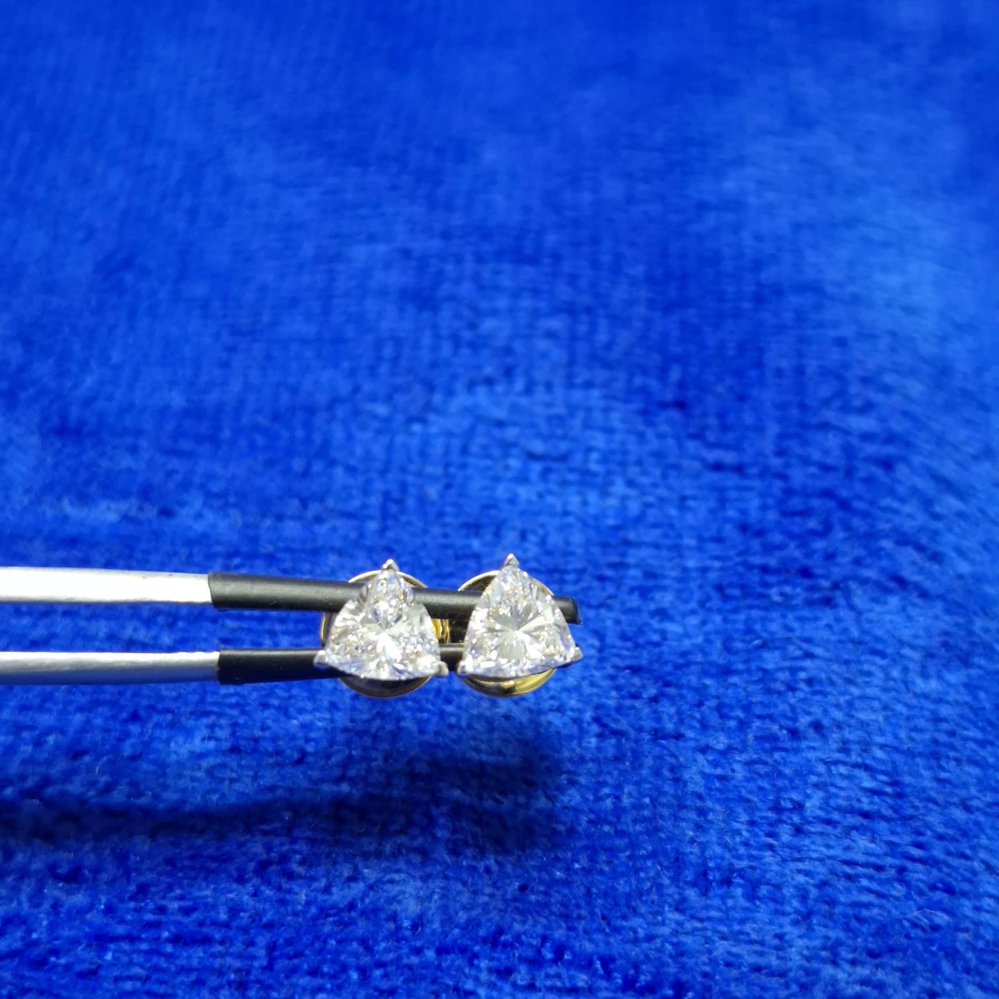 May include: A pair of diamond stud earrings held by tweezers against a blue background. Each earring features a triangular, clear diamond set in a gold base. The diamonds sparkle, reflecting light, and are the focal point of the image. The earrings are presented close-up, highlighting their shape and clarity.