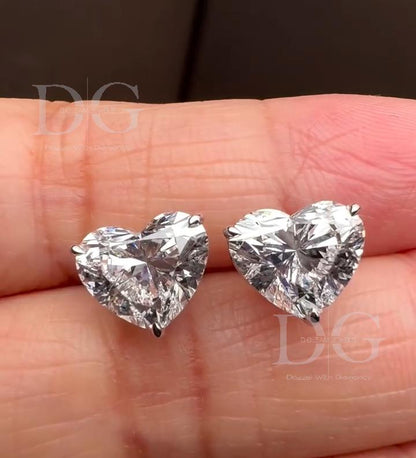 May include: A pair of heart-shaped diamond stud earrings. The earrings are set in a silver metal and are displayed on a light pink background. The diamonds have a brilliant cut, reflecting light with a bright sparkle. The earrings are a classic and elegant jewelry piece.