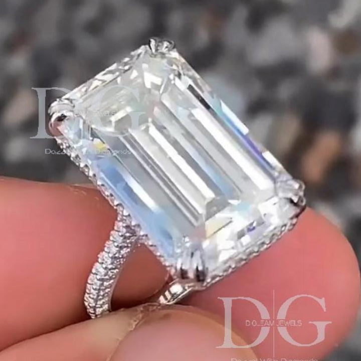 May include: An emerald-cut diamond ring. The large, rectangular diamond is clear and faceted, set on a band with smaller, sparkling stones. The ring is held up, showcasing the intricate details of the setting and the brilliance of the gemstones. The ring is from DO DREAM JEWELS.
