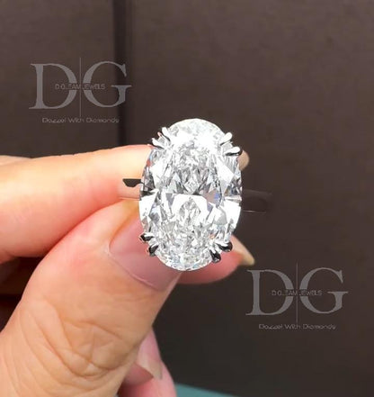 May include: An oval-cut diamond engagement ring. The large, clear diamond is the focal point, set on a silver band with prongs. The ring is held up, showcasing the diamond's facets. The text 'DG DO-FAM JEWELS Dozael With Diamonds' is visible.