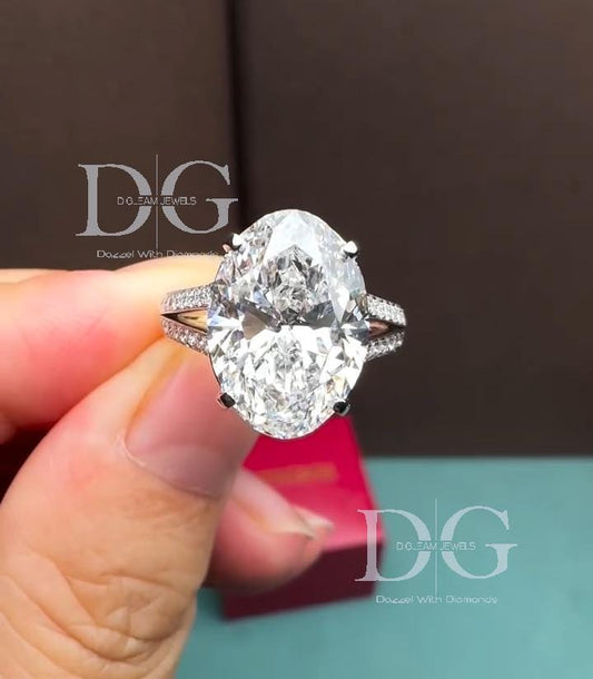May include: An oval-cut diamond engagement ring. The ring features a large, clear oval-shaped diamond as the center stone. The band is adorned with smaller diamonds. The ring is held against a blurred background with the text 'D.G. DREAM JEWELS'.