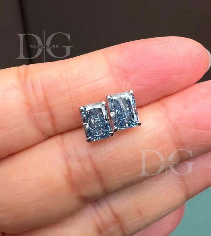 May include: A pair of radiant-cut blue diamond stud earrings. The earrings are set in a silver metal, and each diamond has a rectangular shape with cut corners. The diamonds have a light blue hue and sparkle. The earrings are displayed on a pink surface.