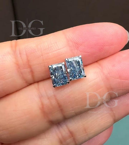 May include: A pair of radiant-cut blue diamond stud earrings. The earrings are set in a silver metal, and each diamond has a rectangular shape with cut corners. The diamonds have a light blue hue and sparkle. The earrings are displayed on a pink surface.