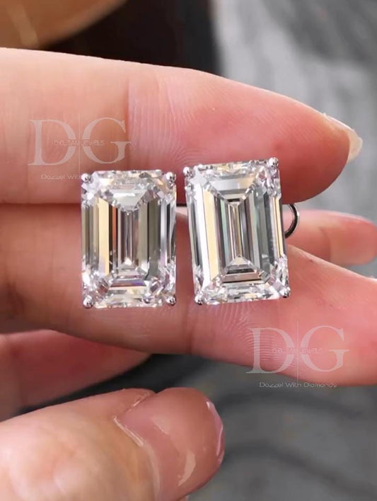 May include: A pair of emerald-cut diamond stud earrings. Each earring features a rectangular, clear diamond with multiple facets, set in a silver-colored metal. The earrings are held against a blurred background, with the text 'DG Dream Jewels Dazzle With Diamonds' visible.