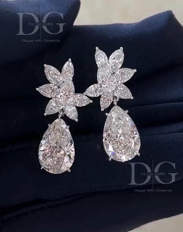 May include: A pair of diamond earrings. Each earring features a floral cluster of small, clear diamonds above a large, pear-shaped diamond. The earrings are set in a silver metal and rest on a dark blue surface. The brand name 'DG DOZEAM JEWELS Dazzle With Diamonds' is visible.