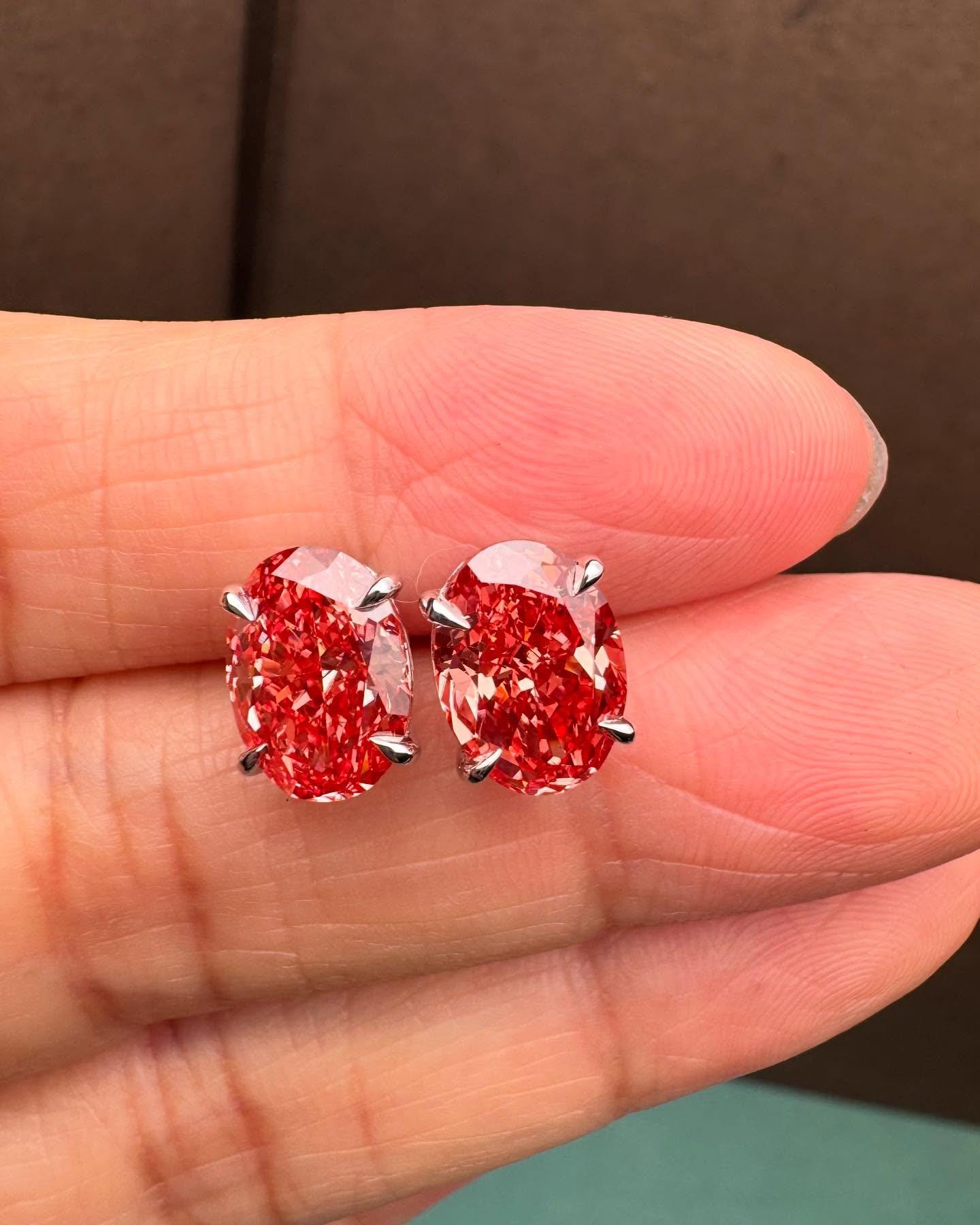 May include: A pair of oval-shaped red gemstone earrings. Each earring features a vibrant, deep red stone held in place by four silver prongs. The stones have a faceted appearance, catching the light and creating a sparkling effect. The earrings are set against a neutral background.