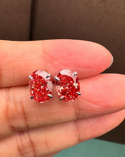 May include: A pair of oval-shaped red gemstone earrings. Each earring features a vibrant, deep red stone held in place by four silver prongs. The stones have a faceted appearance, catching the light and creating a sparkling effect. The earrings are set against a neutral background.